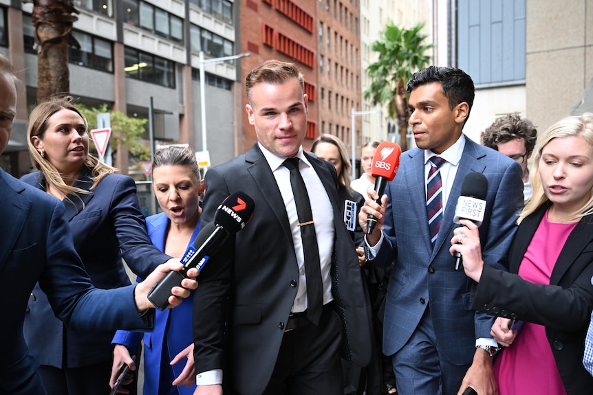 A young man dressed in a suit and tie arrives at a court surrounded by media