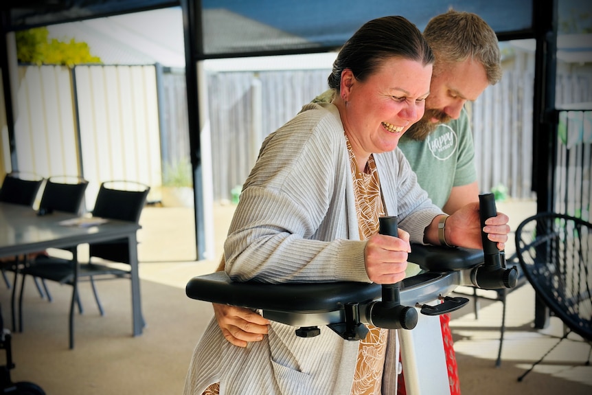 A woman smiling and holding a walking aid by being assisted by her physio