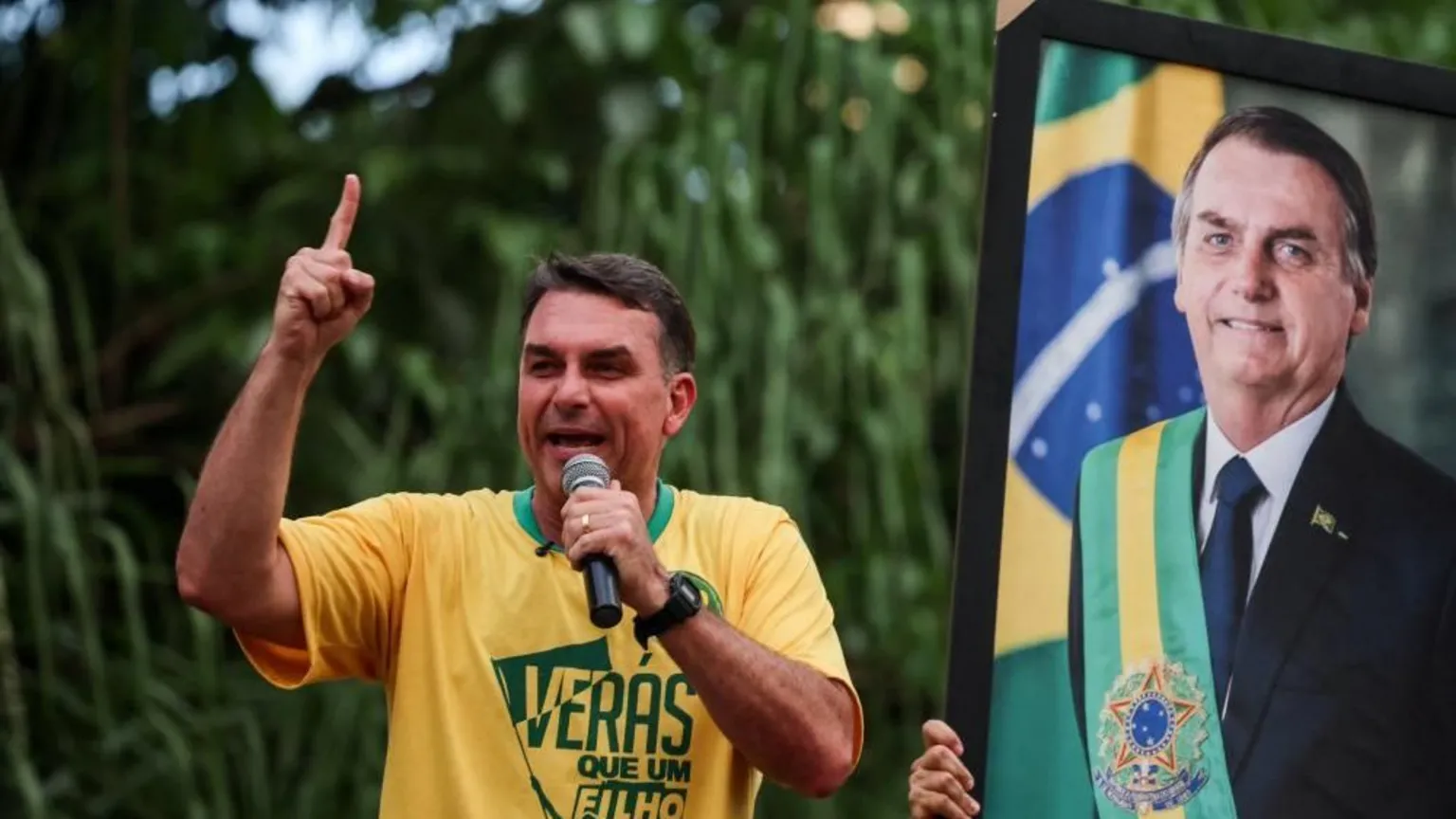 Adriano Machado/Reuters Brazilian presidential candidate Flavio Bolsonaro of the Liberal Party (PL) speaks next to a portrait of his father, Brazil's former President Jair Bolsonaro, during a campaign rally in Goiania, Brazil, September 27, 2026. He is wearing a yellow T0shirt and holds up his fight index finger as he speaks into a microphone.