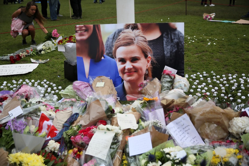 Floral tributes scattered around a large printed photograph of a woman resting on the lawn outside parliament.